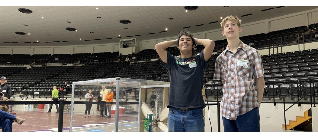 Two students looking at camera with wind turbine in wind tunnel behind them